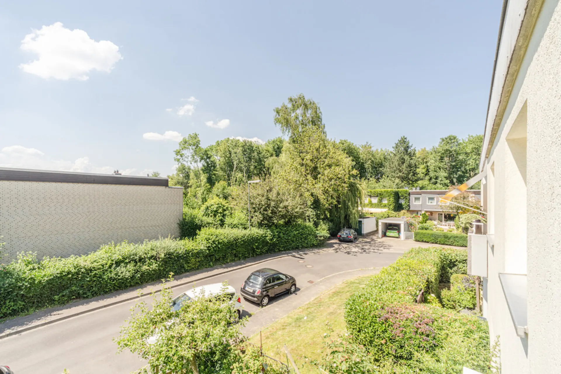 Blick auf eine ruhige Wohnstraße mit geparkten Autos, Häusern und viel Grün bei sonnigem Wetter.