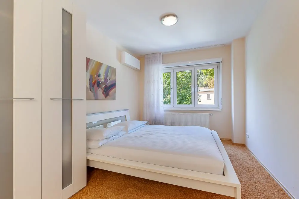 Helles Schlafzimmer mit weißem Bett, Kleiderschrank, Fenster und abstraktem Bild an der Wand.