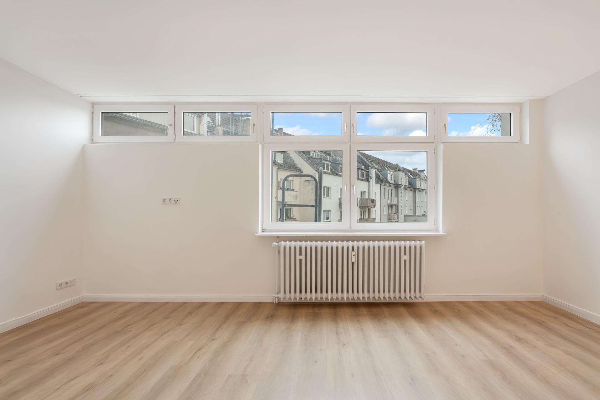 Leerer Raum mit hellem Holzfußboden, weißen Wänden und großem Fenster mit Heizkörper.