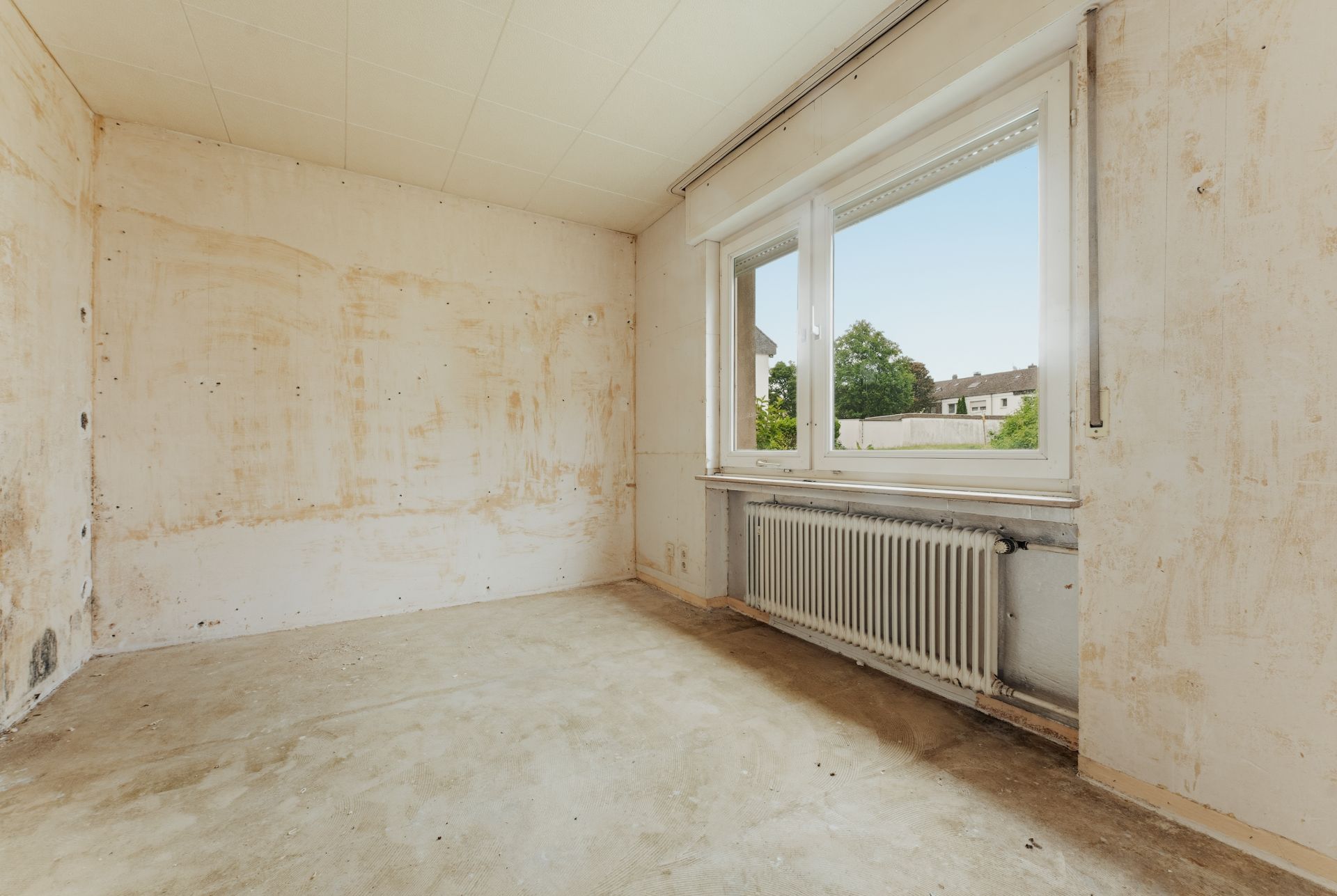 Leerer Raum mit abgenutzten Wänden, großem Fenster und Heizkörper unter dem Fenster.