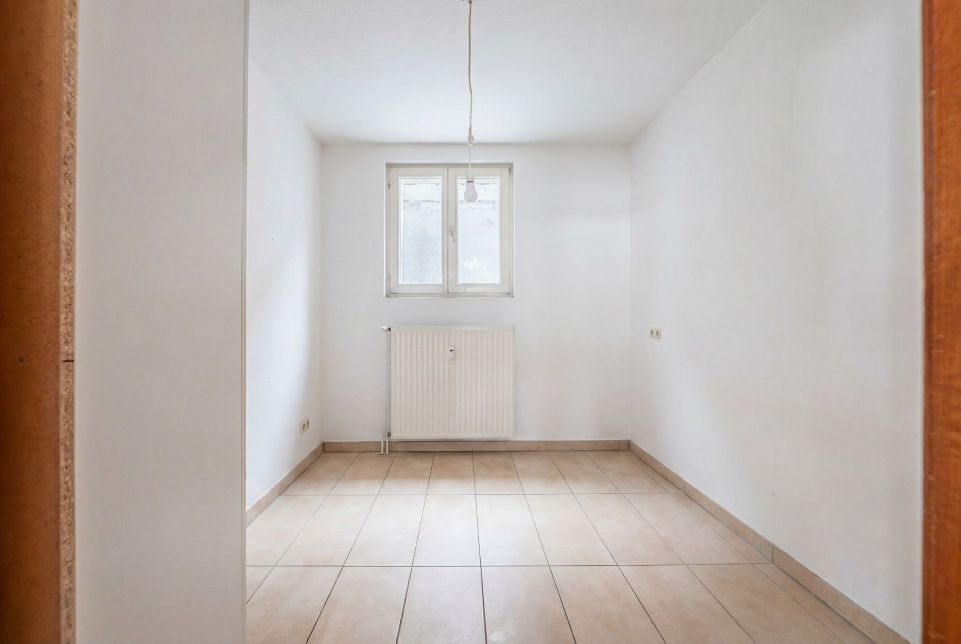 Leerer Raum mit weißen Wänden, Fliesenboden, Fenster und Heizkörper.