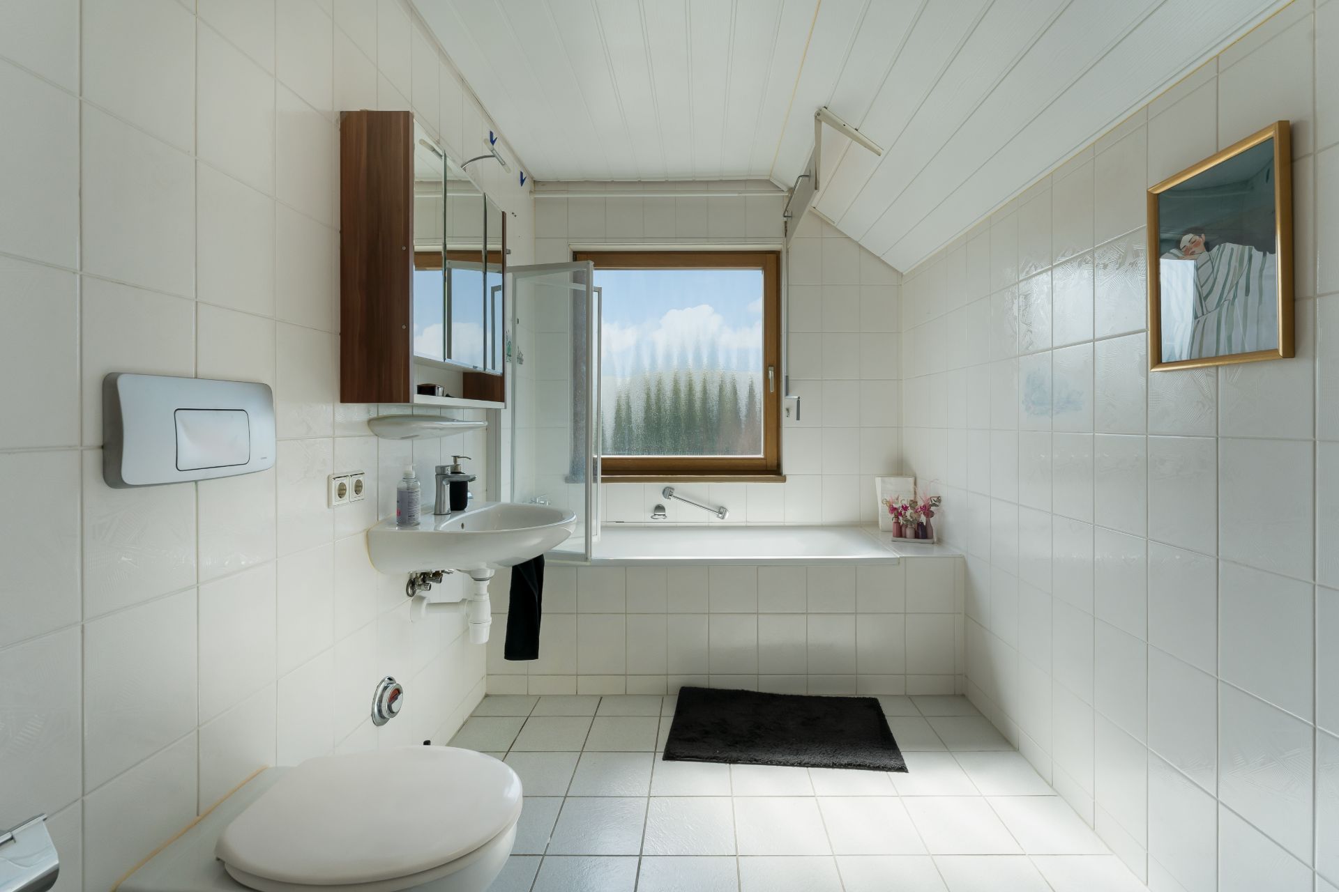 Helles Badezimmer mit Badewanne, Fenster, Waschbecken und Toilette, weiße Fliesen und Wandbild.