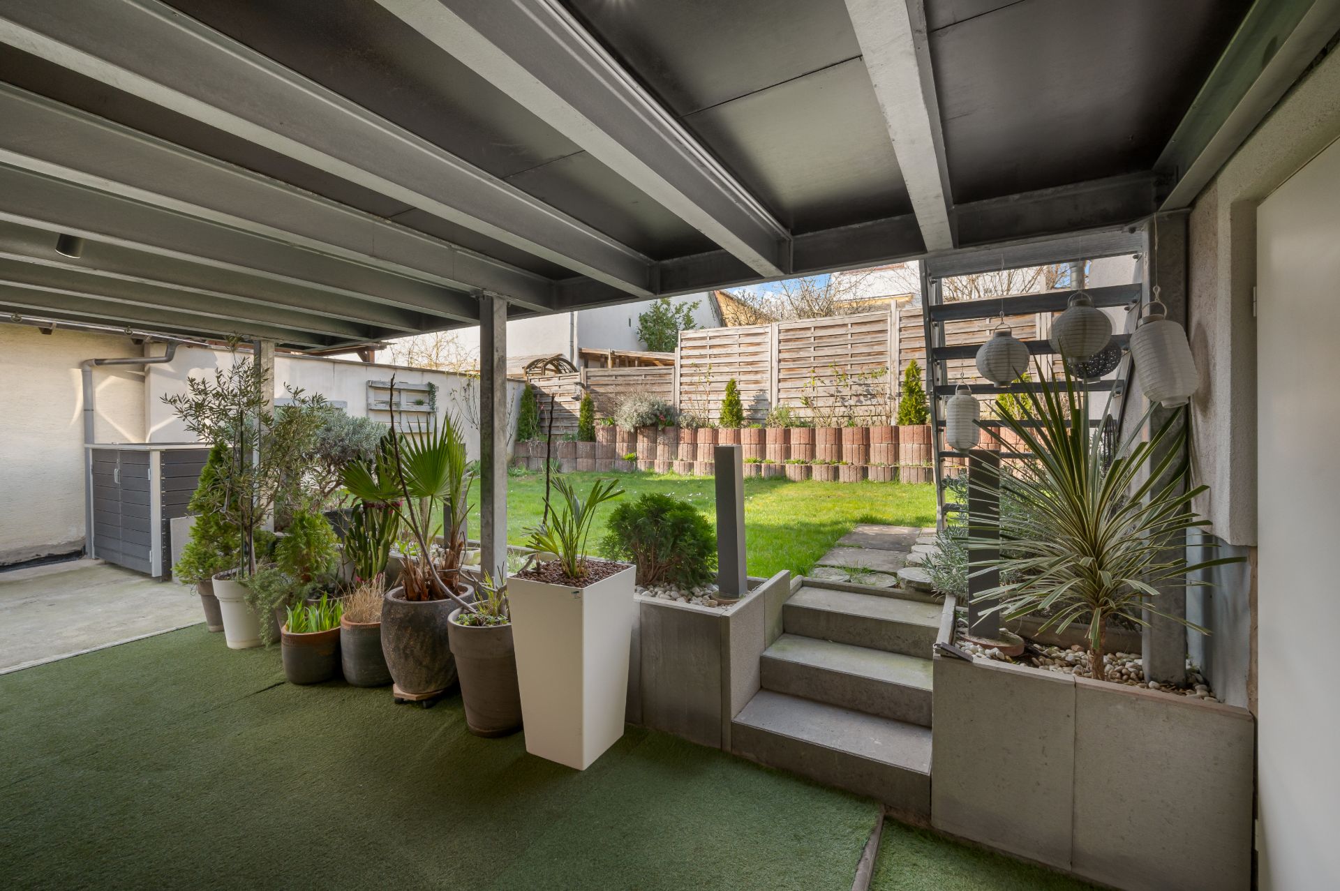 Überdachte Terrasse mit Pflanzen und Treppe zu einem Garten mit Rasen und Holzzaun.