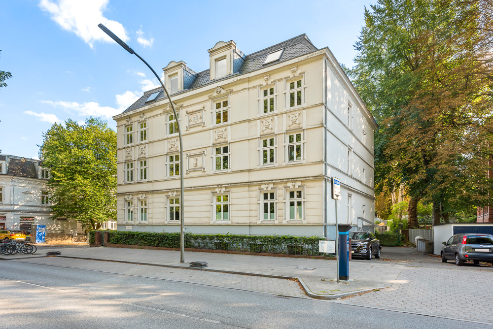 Weiße Wohnhausfassade mit vielen Fenstern an einer Straße, Bäume im Hintergrund.