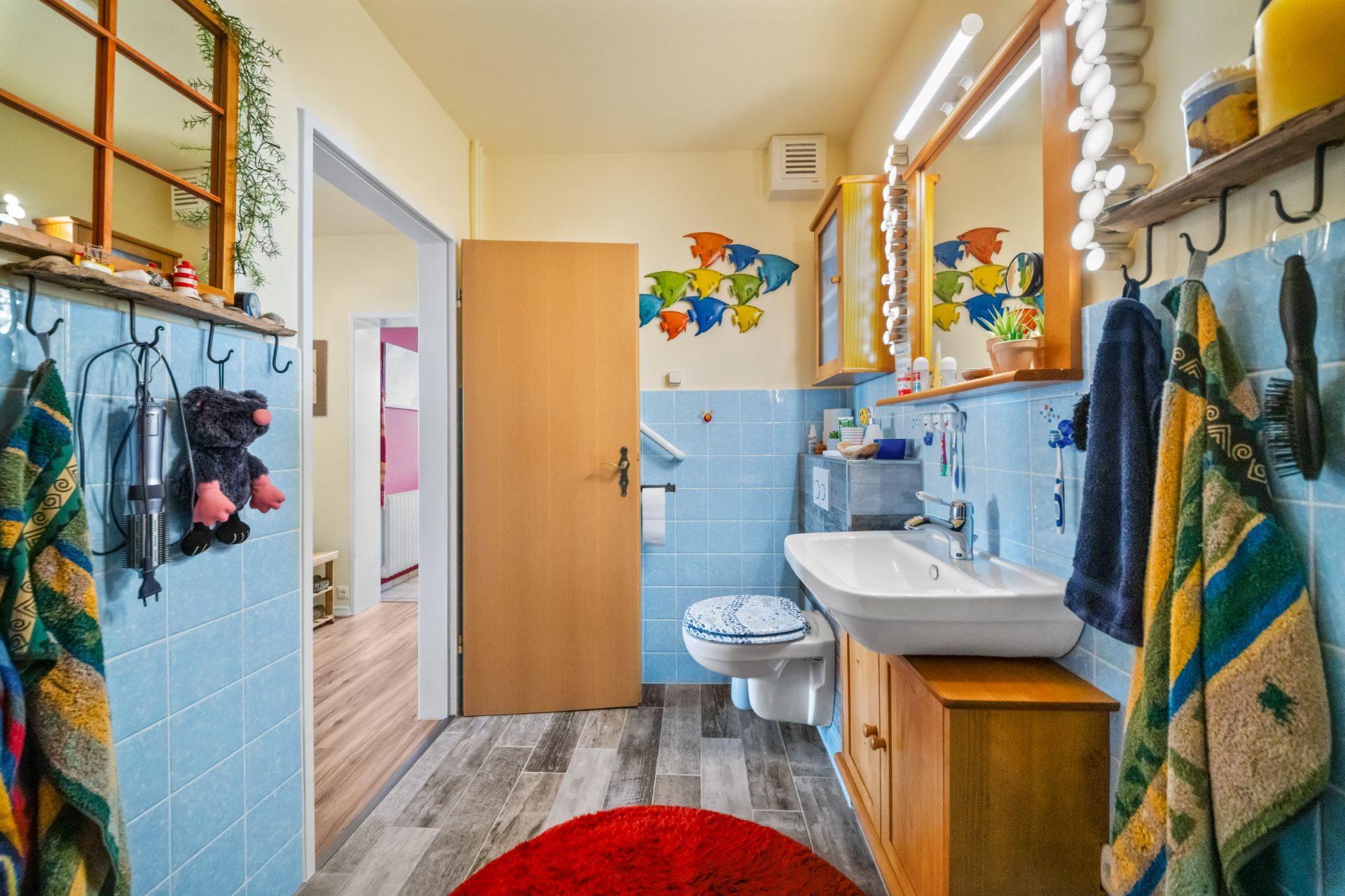 Badezimmer mit blauen Fliesen, Waschbecken, Spiegel, WC, bunten Handtüchern und Wanddeko mit Fischen.