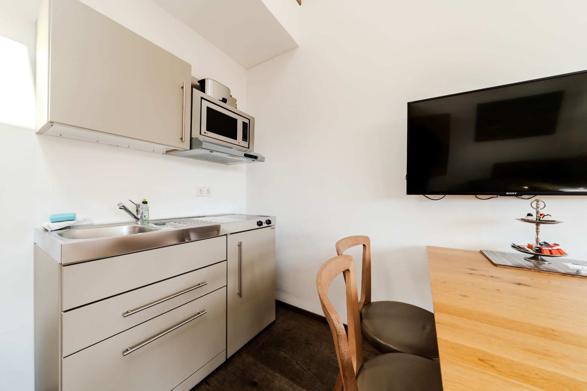 Kleine Küche mit Spüle, Herd, Mikrowelle, Esstisch, Stühlen und einem Fernseher an der Wand.