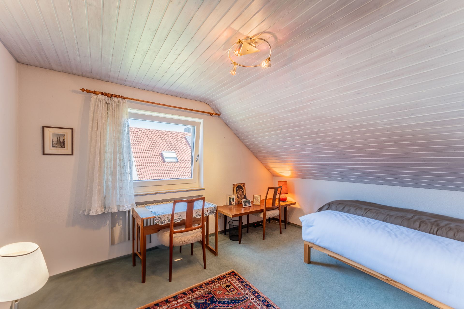 Helles Dachzimmer mit Bett, zwei Schreibtischen, Stuhl und Teppich, Fenster mit Gardine.