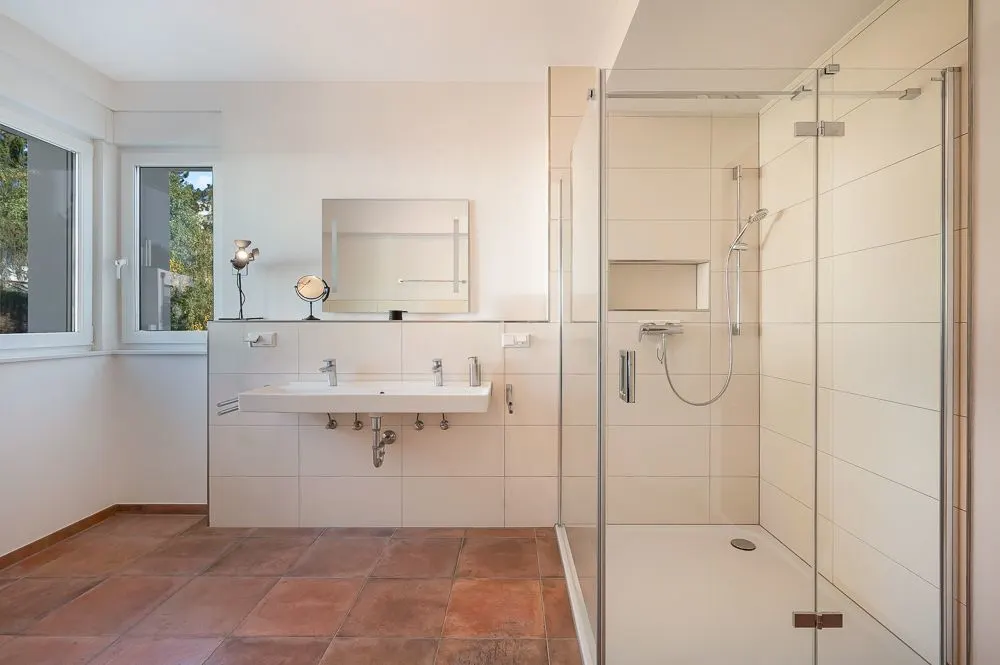 Modernes Badezimmer mit großer Dusche, zwei Waschbecken und Spiegel, Fliesenboden und zwei Fenstern.