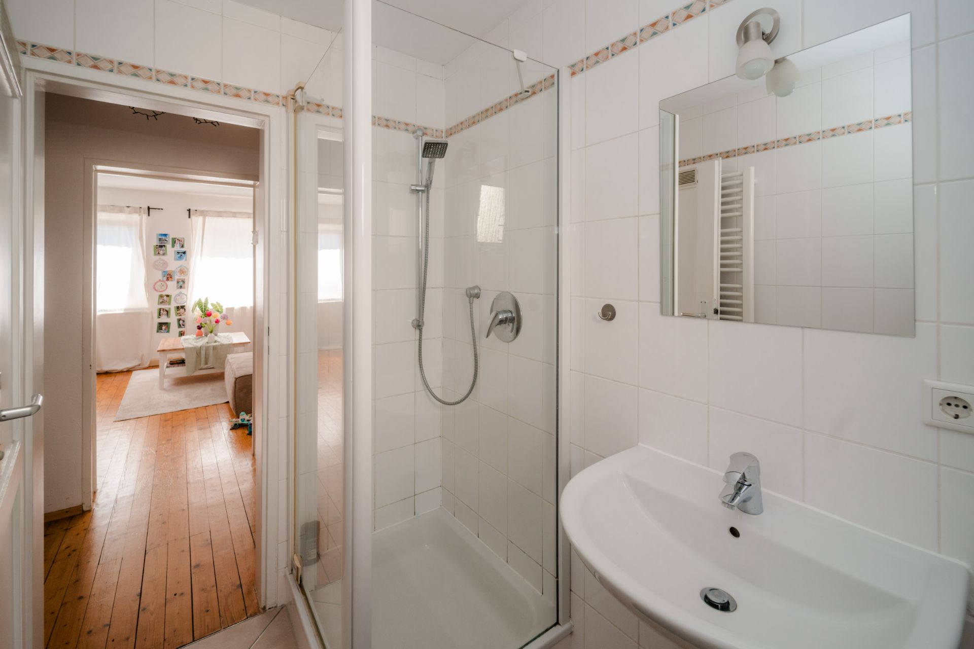 Kleines Badezimmer mit Dusche, Waschbecken und Spiegel, Blick ins helle Wohnzimmer mit Holzboden.