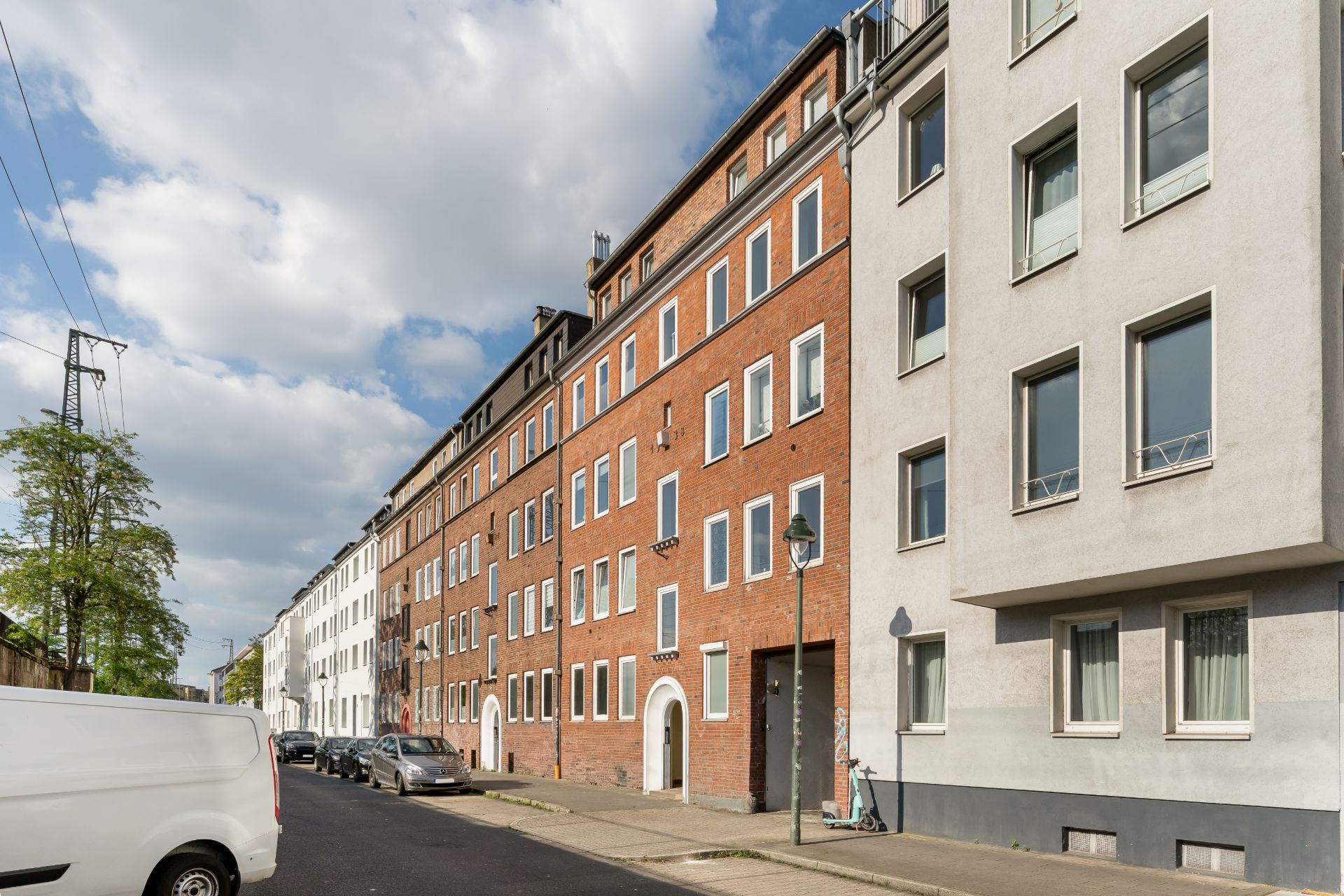 Mehrstöckige Wohnhäuser an einer ruhigen Straße mit geparkten Autos und einem weißen Lieferwagen.