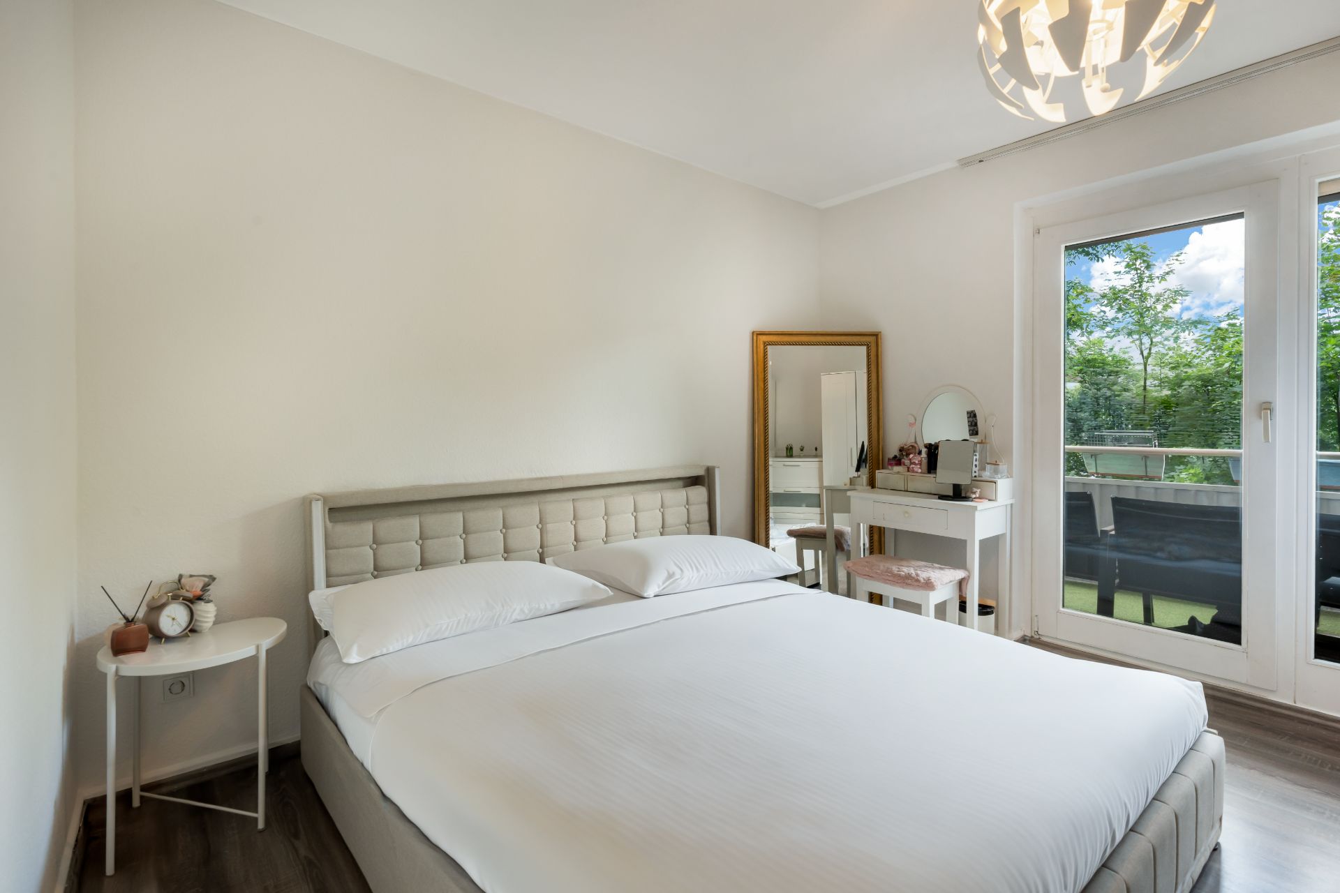 Helles Schlafzimmer mit Doppelbett, Schminktisch, großem Fenster und Balkonblick.