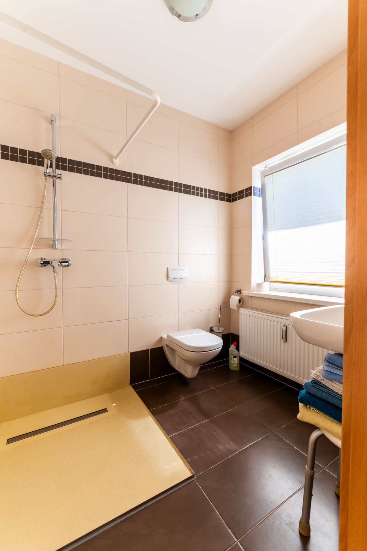 Barrierefreies Badezimmer mit Dusche, Wand-WC, Waschbecken und Fenster mit Plissee.