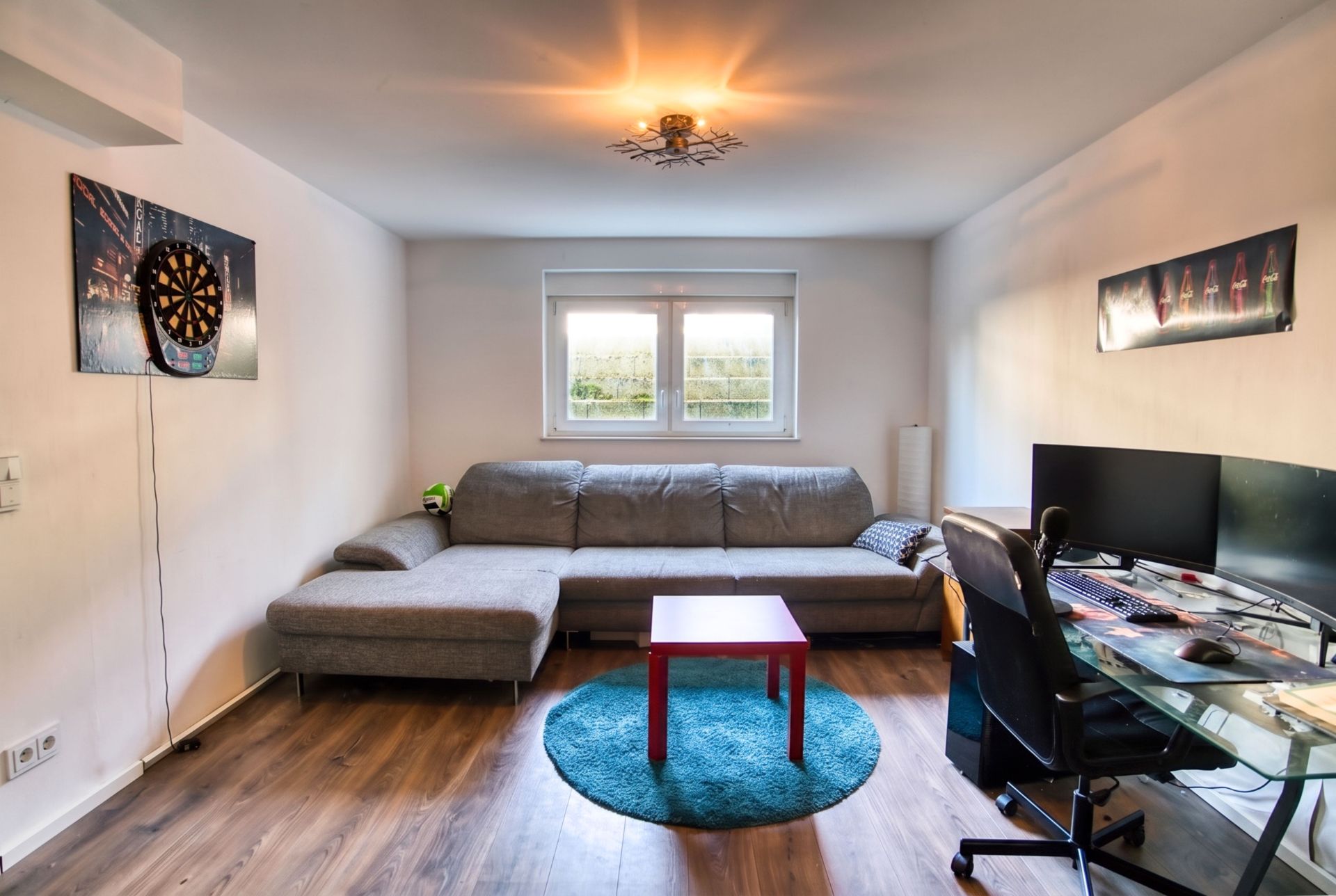 Wohnzimmer mit grauem Sofa, Schreibtisch, Computer und Dartscheibe an der Wand.