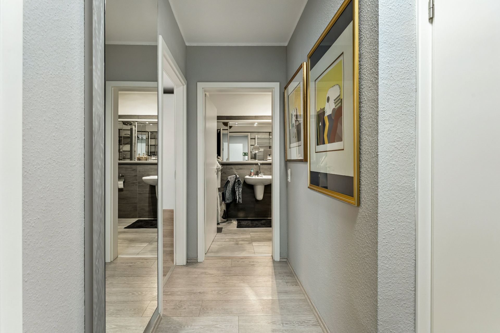 Heller Flur mit Bildern an der Wand und Blick ins Badezimmer mit Spiegel und Waschbecken.