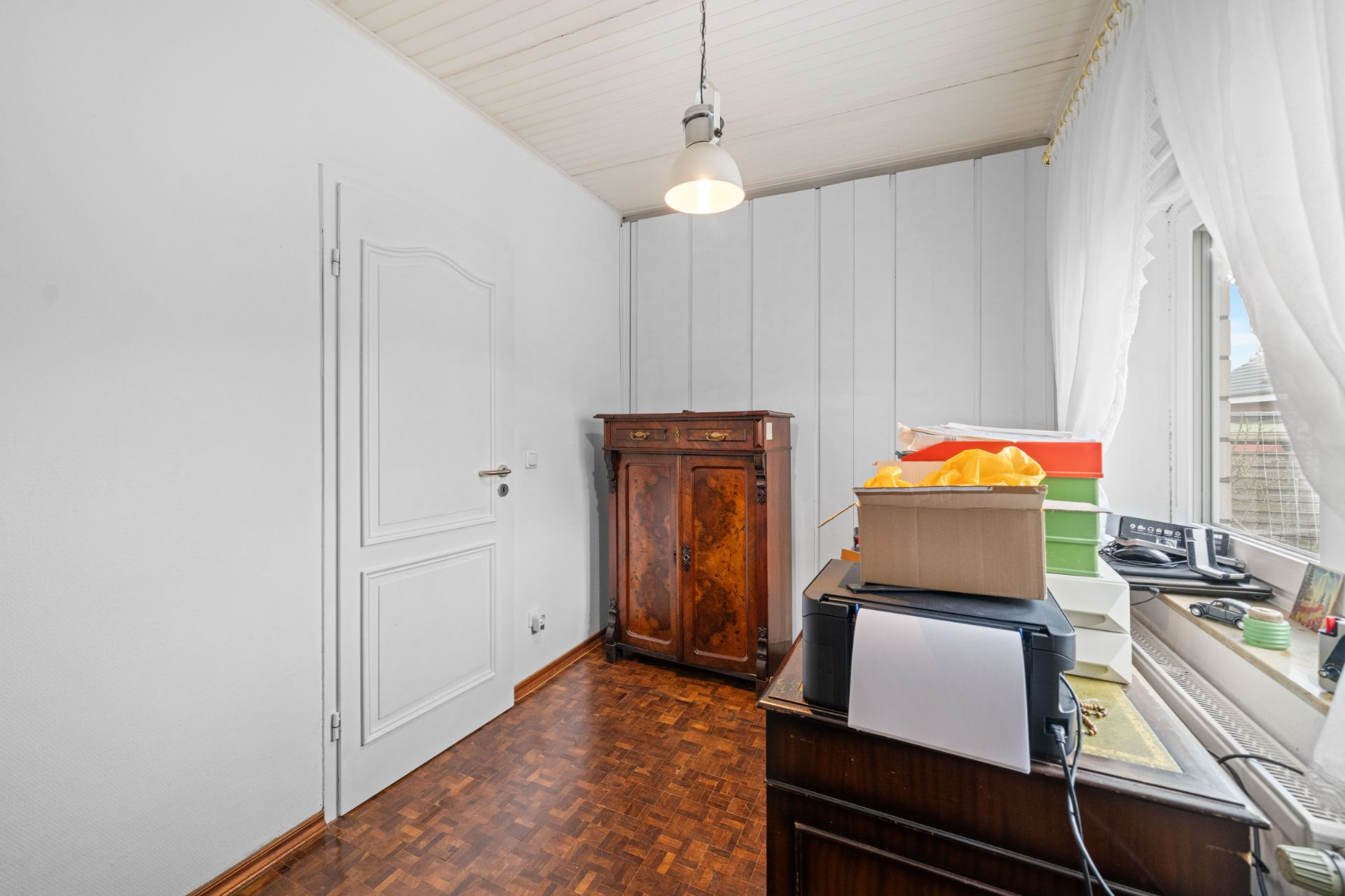 Kleines Zimmer mit Holzboden, Kommode, Schreibtisch, Kartons und Fenster mit weißen Vorhängen.