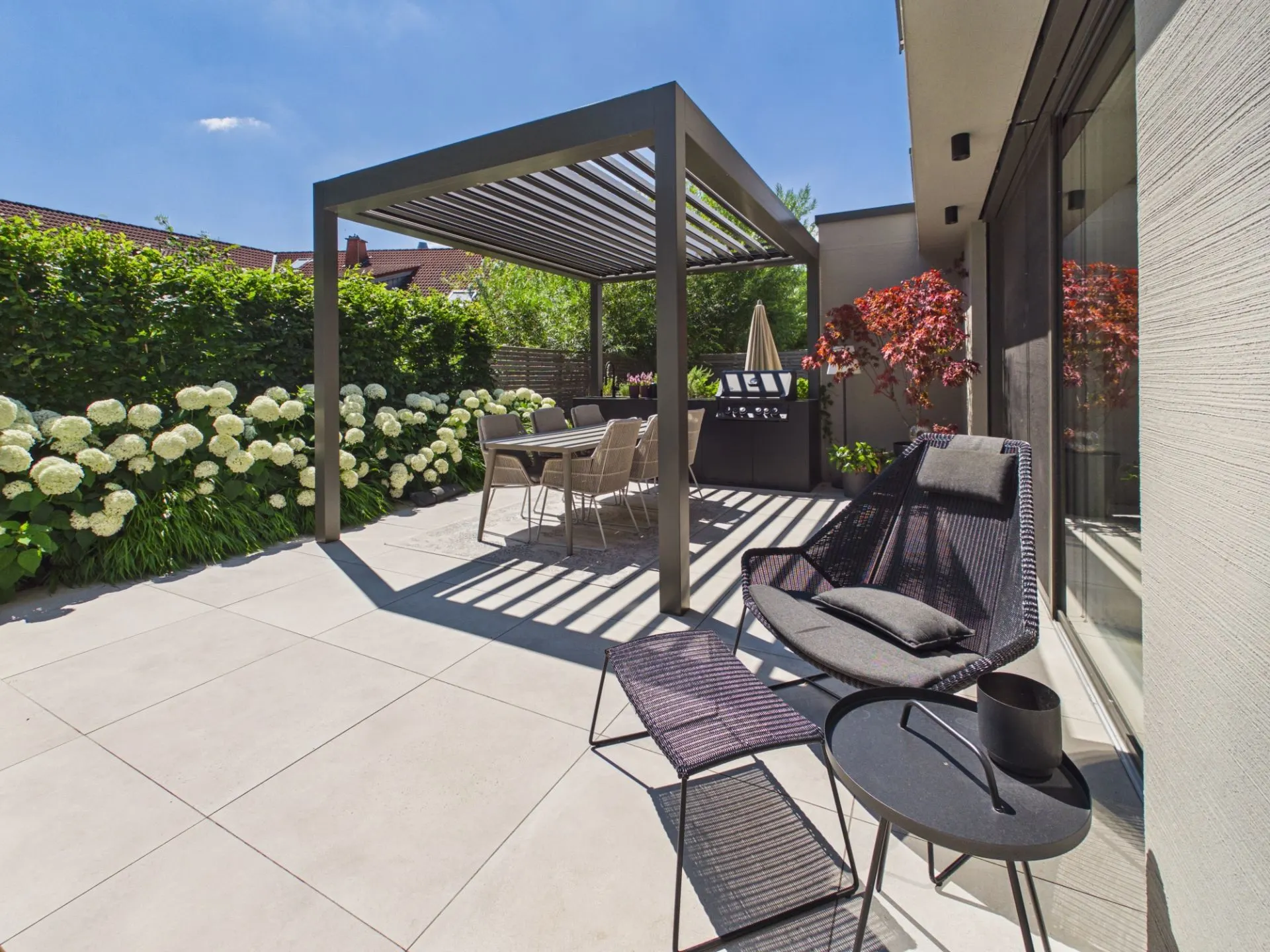 Moderne Terrasse mit Pergola, Sitzbereich, Blumen und Hecke bei sonnigem Wetter.