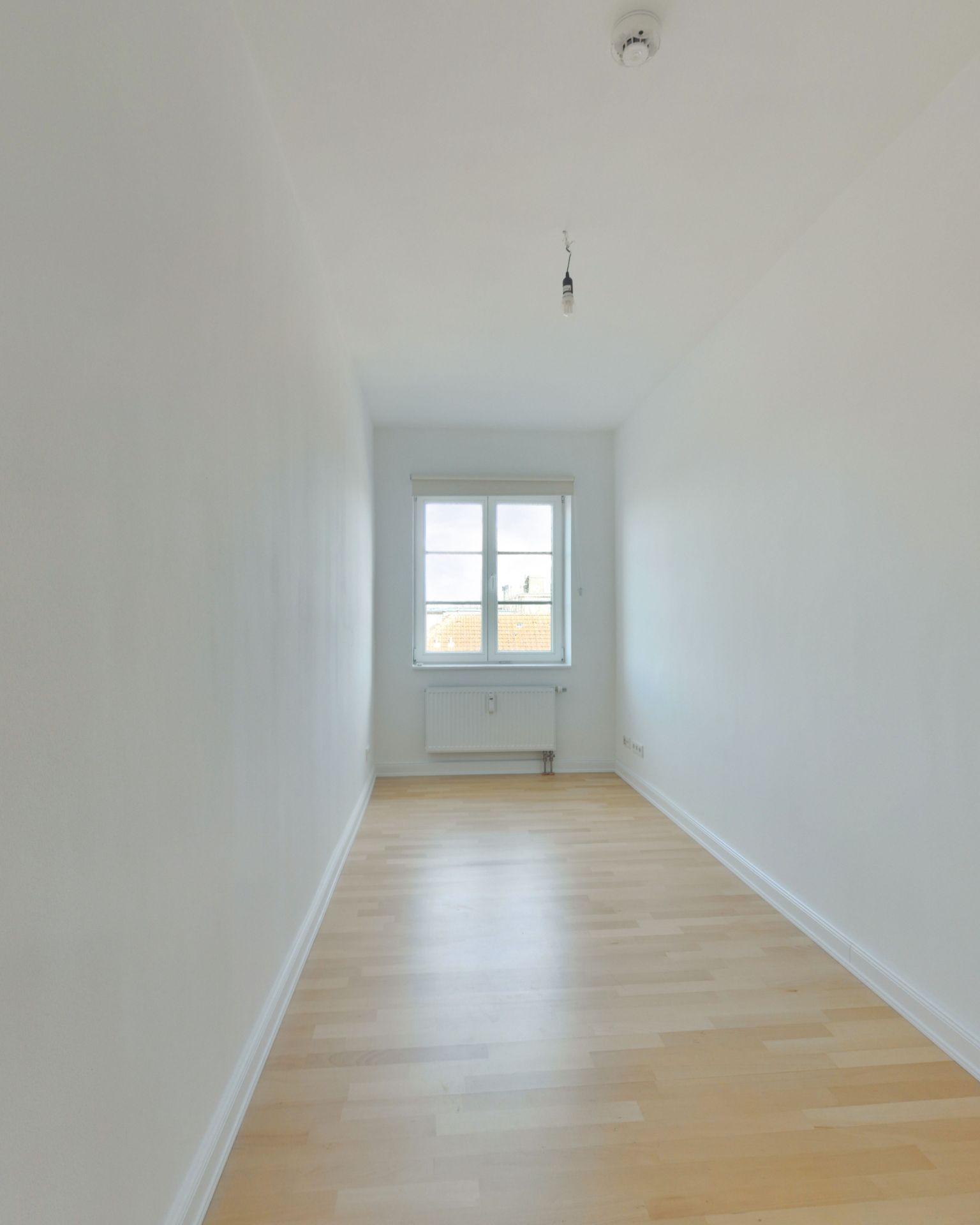 Schmaler, leerer Raum mit hellem Holzboden, weiß gestrichenen Wänden und einem Fenster am Ende.