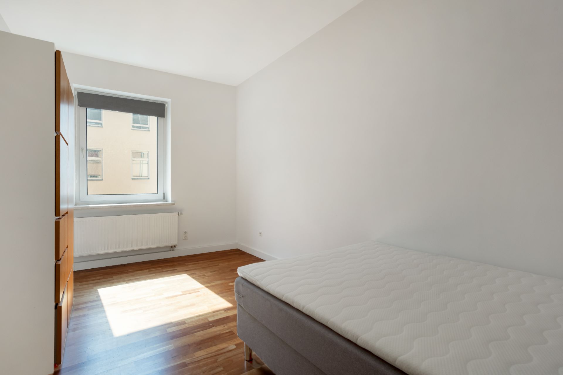 Helles Schlafzimmer mit Fenster, Bett, Holzschrank und Parkettboden.
