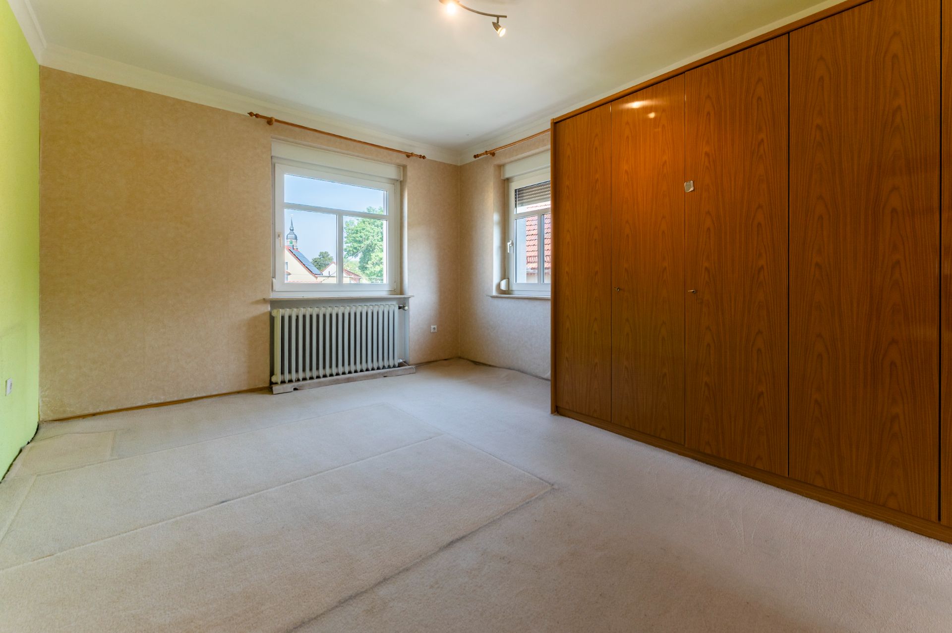 Leeres Zimmer mit Teppichboden, zwei Fenstern und großem Holzschrank an der rechten Wand.