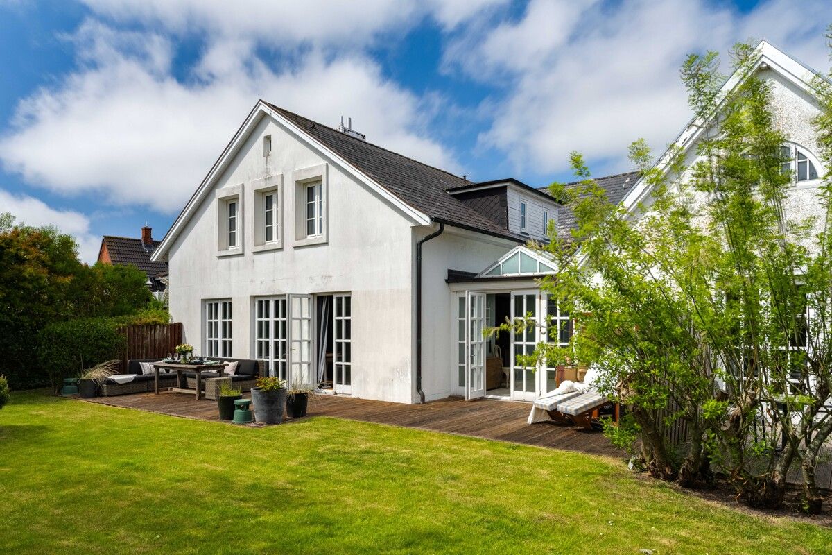 Weißes Einfamilienhaus mit Garten, Holzterrasse und Gartenmöbeln bei blauem Himmel.