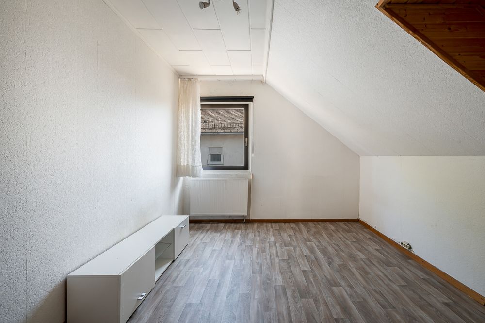 Helles Zimmer mit Dachschräge, Fenster, Vorhang, Heizung und niedrigem Regal auf Holzfußboden.