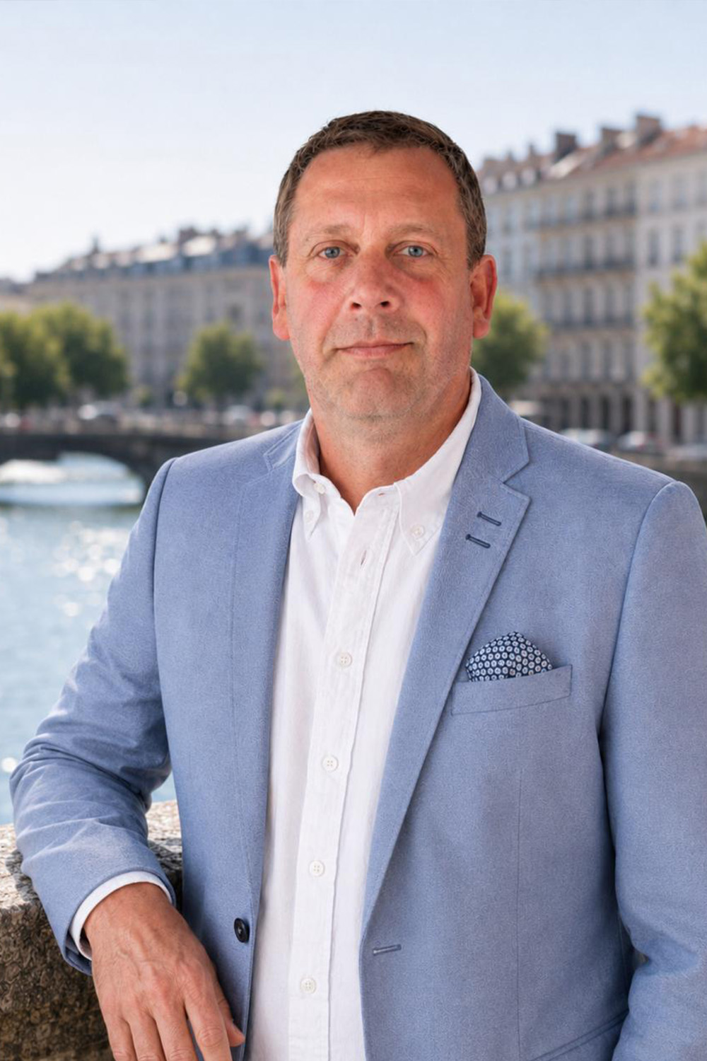 Ein Mann im hellblauen Sakko steht draußen vor einem Fluss, im Hintergrund Wohngebäude.