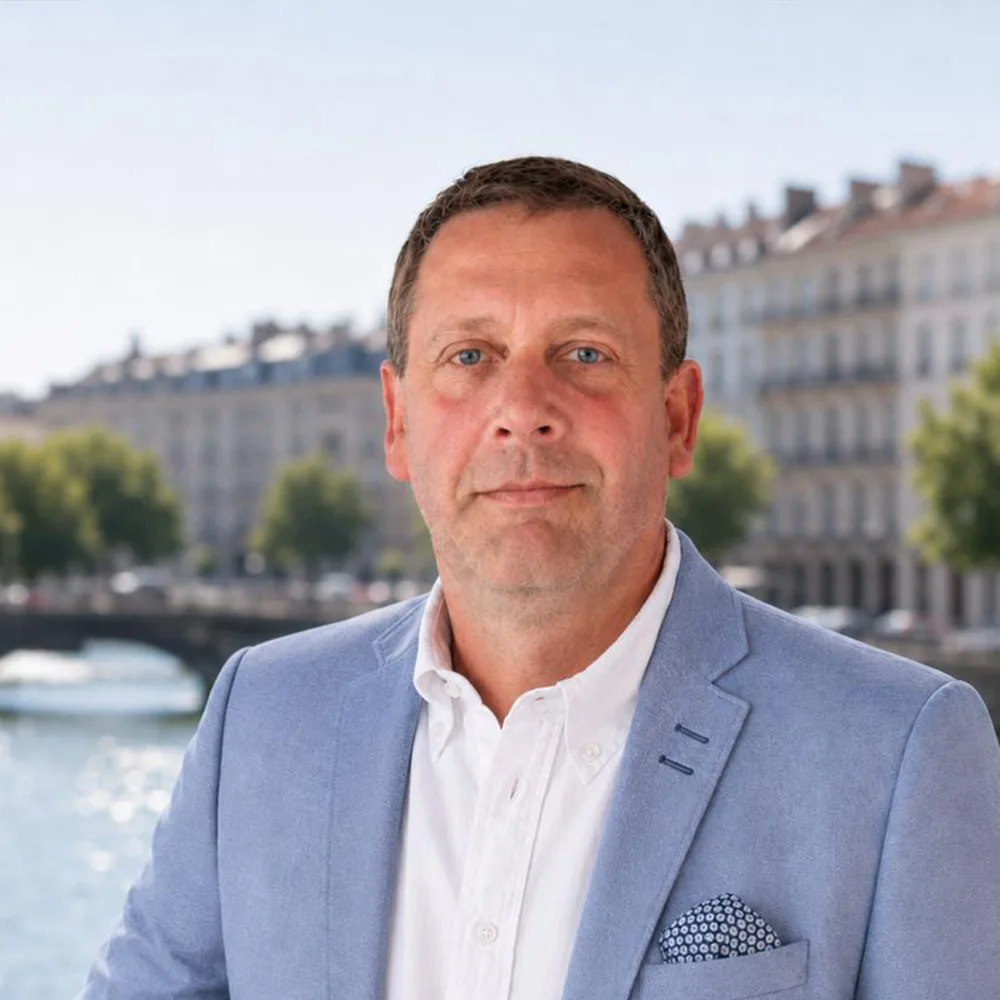 Ein Mann im hellblauen Sakko steht draußen vor einem Fluss, im Hintergrund Wohngebäude.