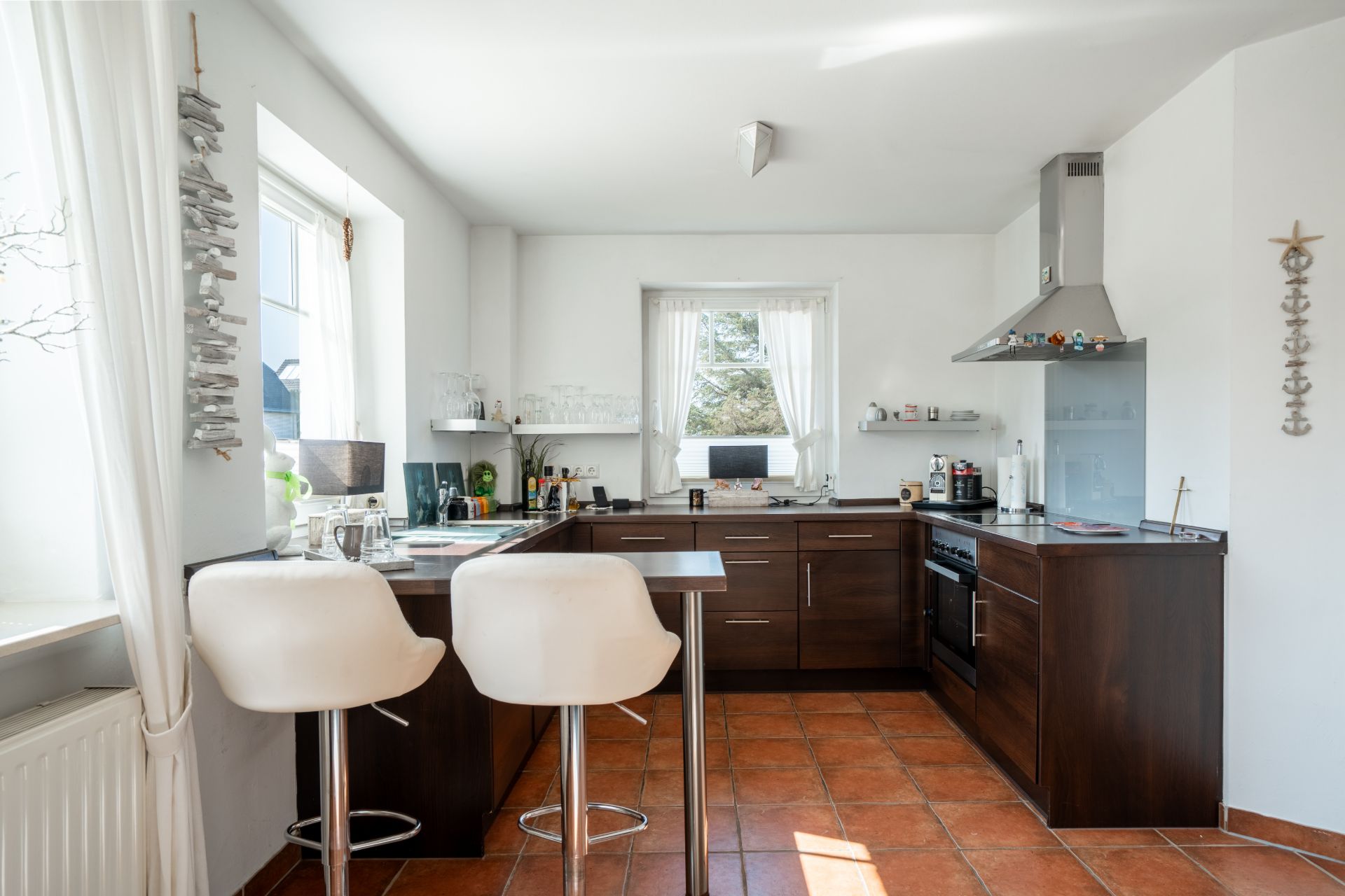 Moderne Küche mit dunklen Schränken, zwei weißen Barhockern und großen Fenstern mit weißen Vorhängen.
