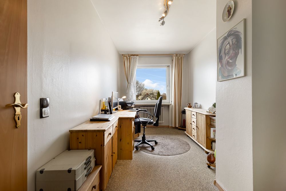 Helles Arbeitszimmer mit Schreibtisch, Stuhl, Regalen und Fenster mit Vorhängen.