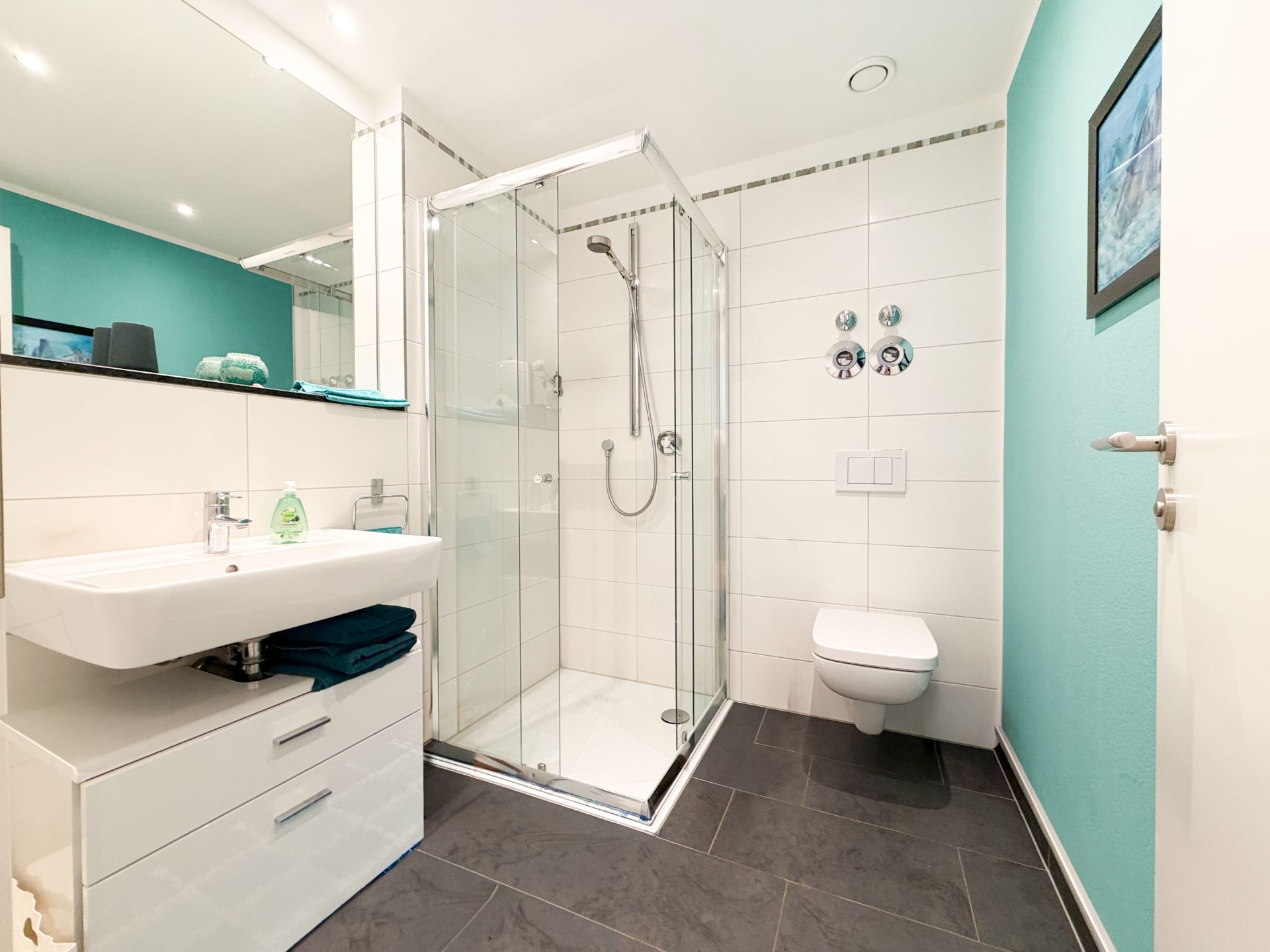 Modernes Badezimmer mit Dusche, Wandwaschbecken und WC, türkisfarbene Wand und Spiegel.