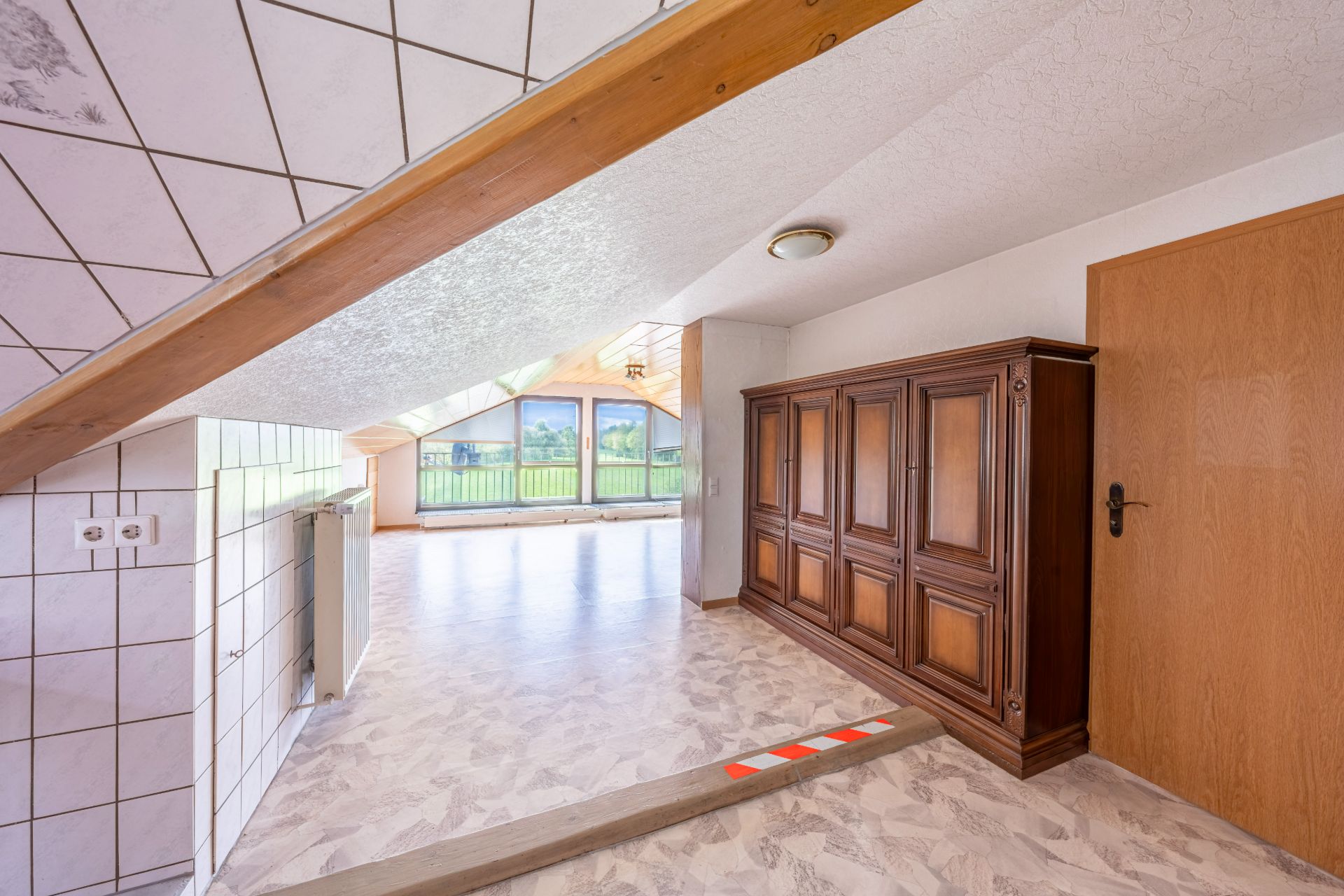 Wohnbereich mit Holzschrank, Dachschräge, großer Fensterfront und heller Bodenfliese.