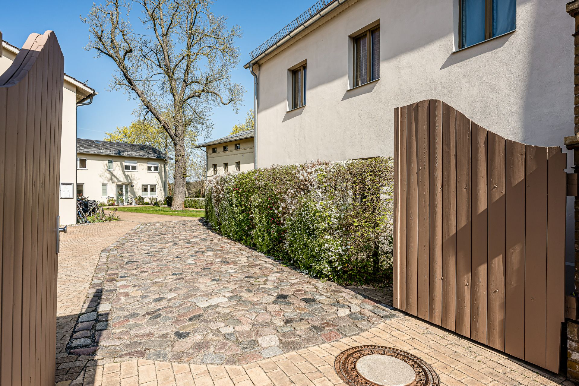 Offenes Holztor mit Pflasterweg, Hecke und Wohnhäusern im Hintergrund.