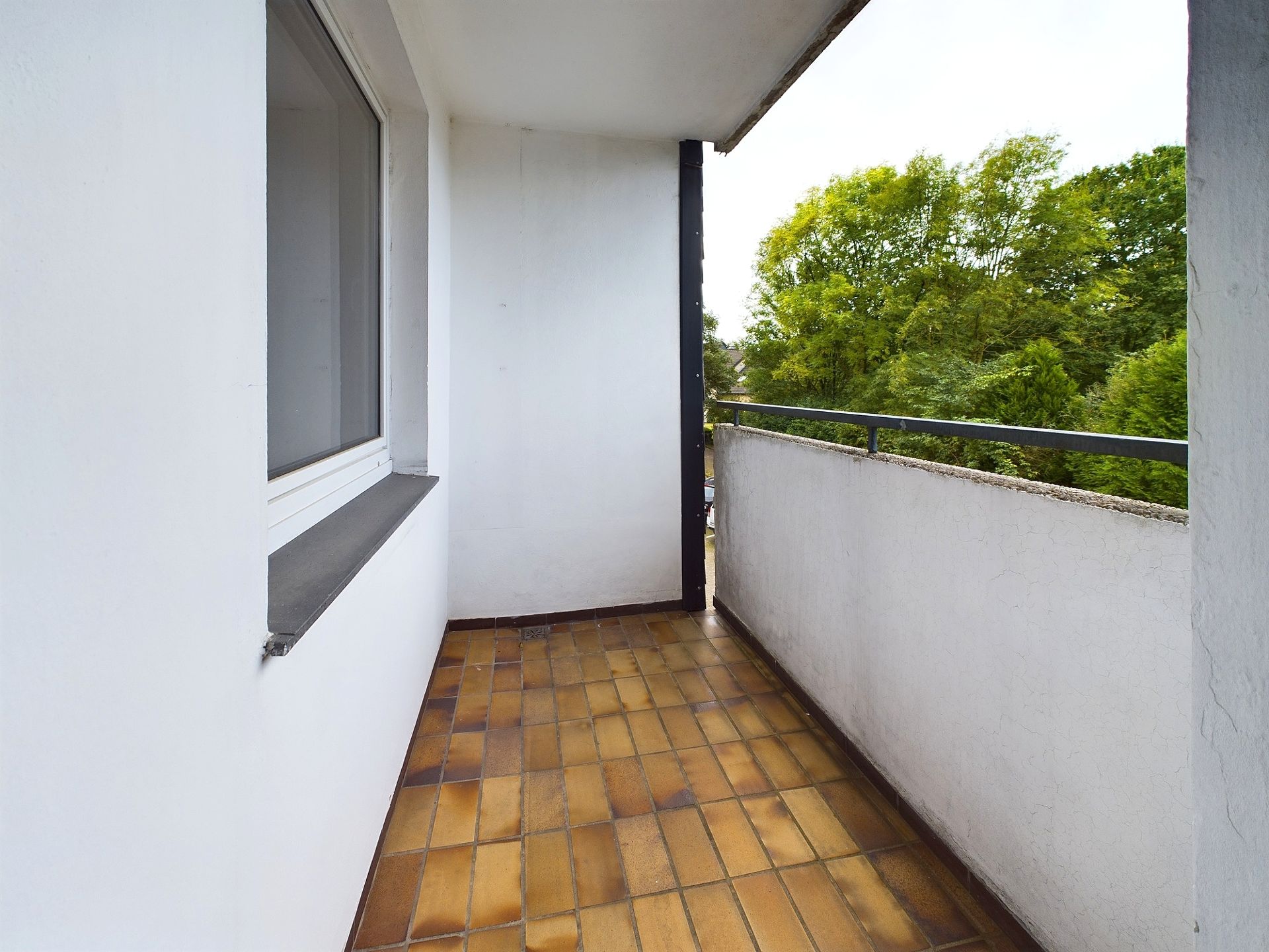 Balkon mit braunen Fliesen, weißer Wand und Blick auf grüne Bäume.