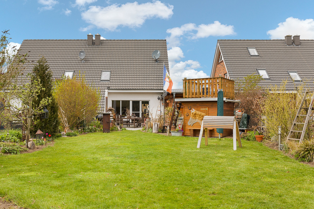 Garten mit Rasen, Spielhaus aus Holz, Sitzbereich und Büschen vor einem Wohnhaus.