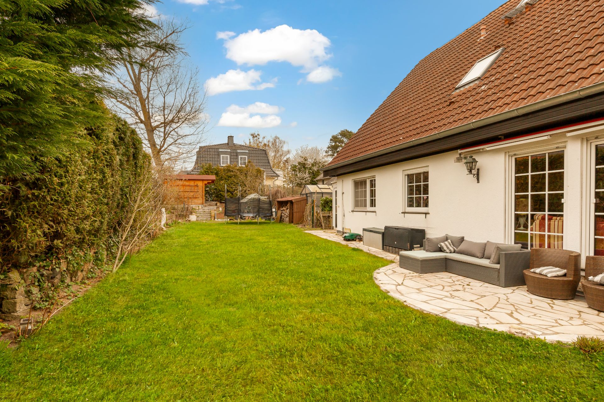 Garten mit Rasen und Terrasse, Gartenmöbeln und Haus mit großen Fenstern.