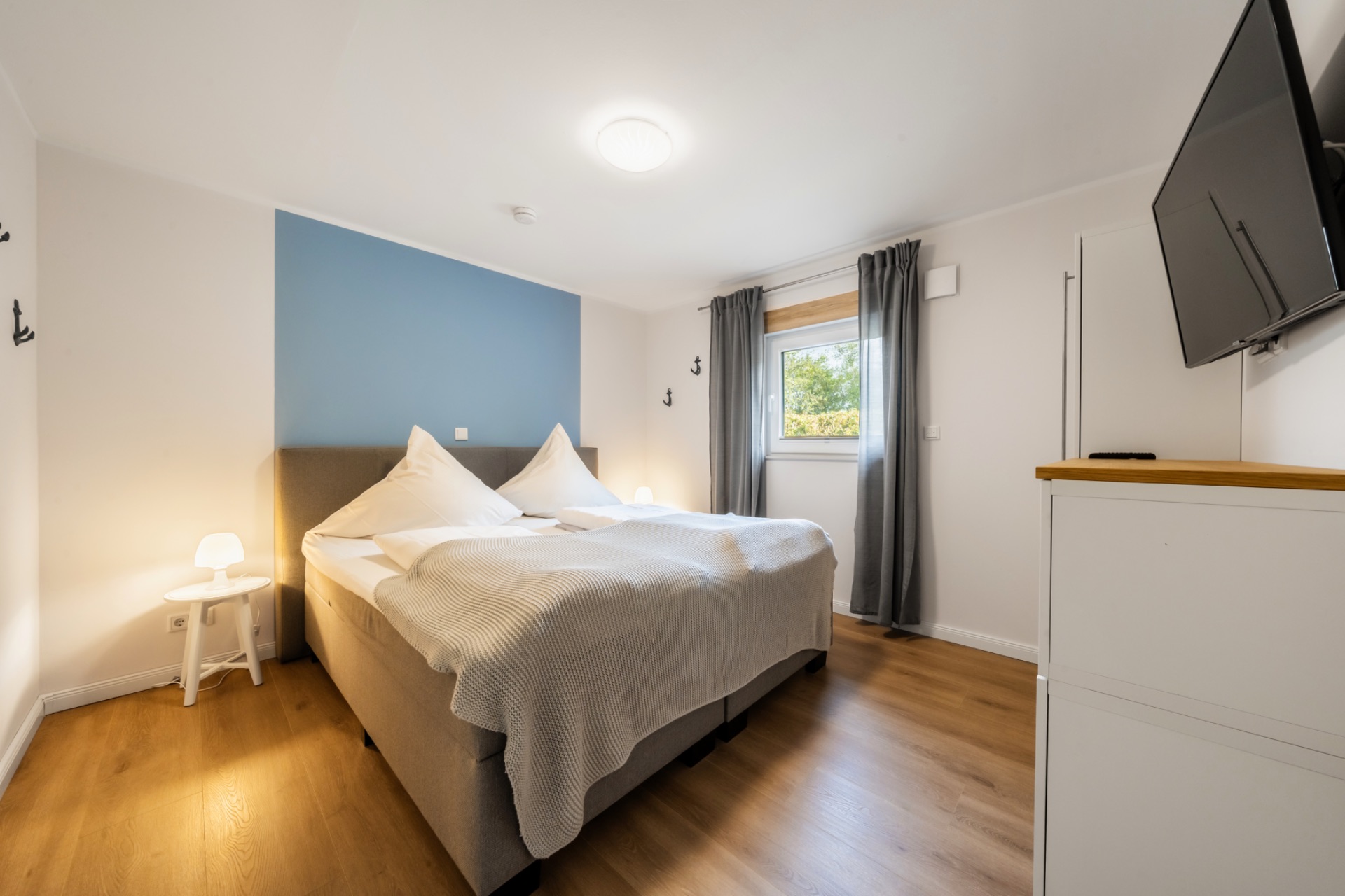 Helles Schlafzimmer mit Doppelbett, Nachttischen, Fenster mit grauen Vorhängen und Wandfernseher.