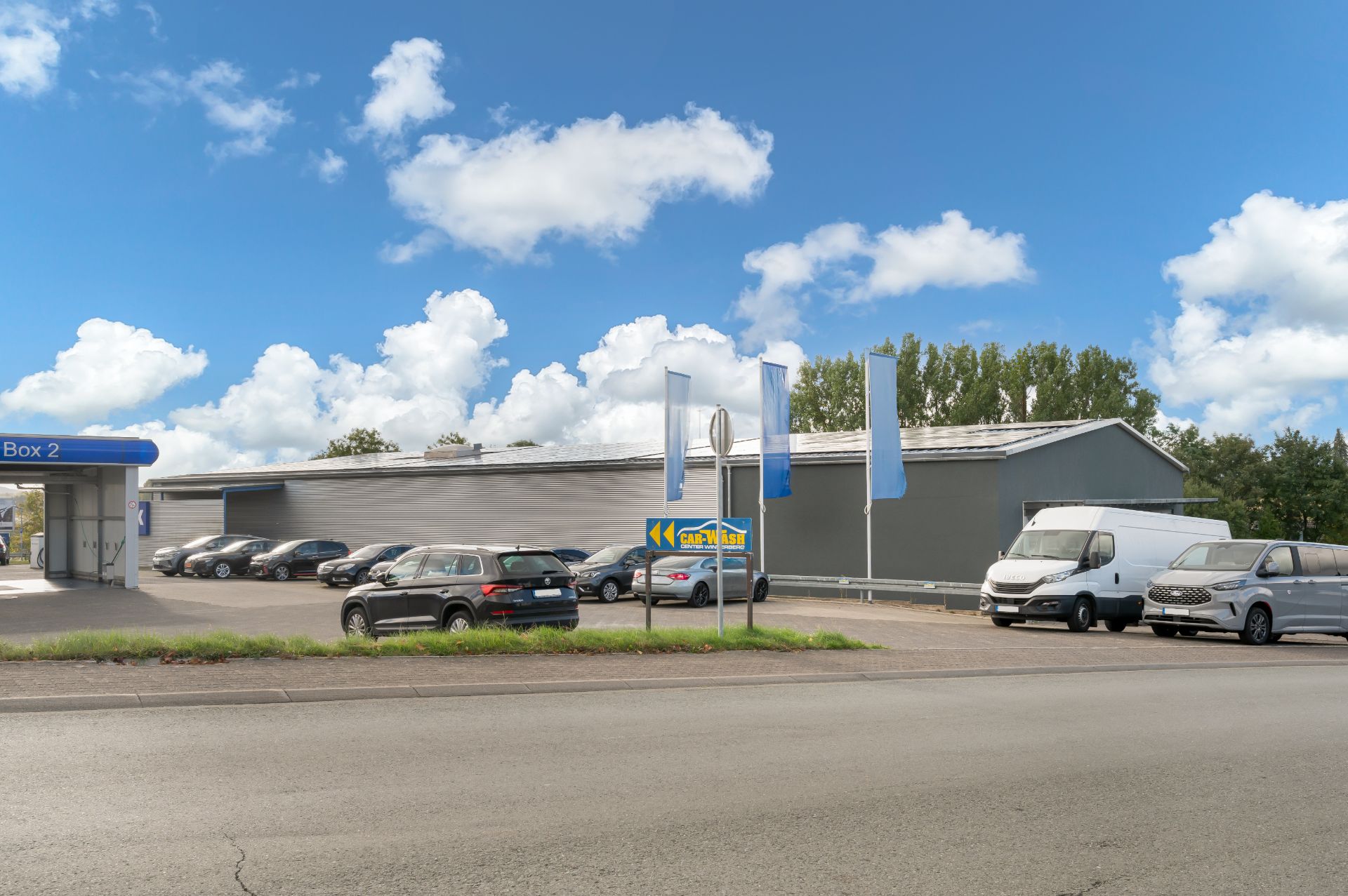 Großer Parkplatz mit mehreren Autos und weißen Fahnen vor einem grauen Gebäude.