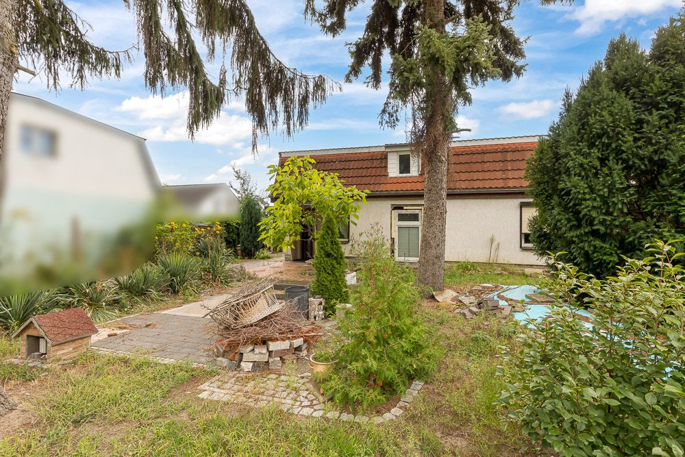 Garten mit Bäumen, Sträuchern, Gehweg und Blick auf ein Haus mit rotem Dach.