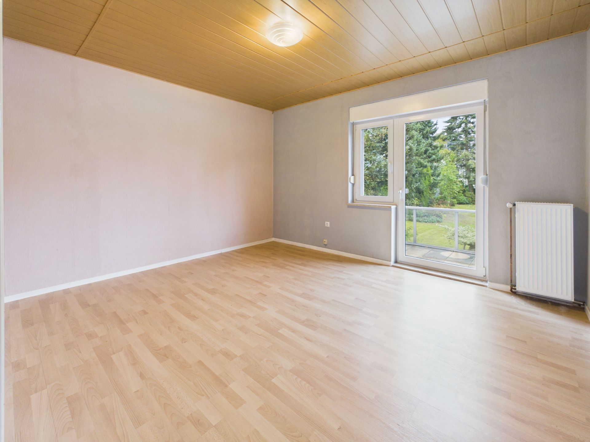 Leeres Zimmer mit Holzdecke, Parkettboden, Fenster und Tür zum Balkon, Blick ins Grüne.