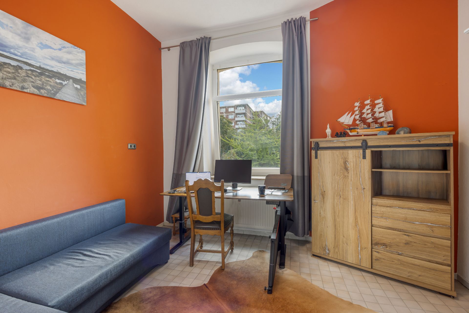 Arbeitszimmer mit orangefarbenen Wänden, Sofa, Schreibtisch, Computer und einem Fenster mit Vorhängen.