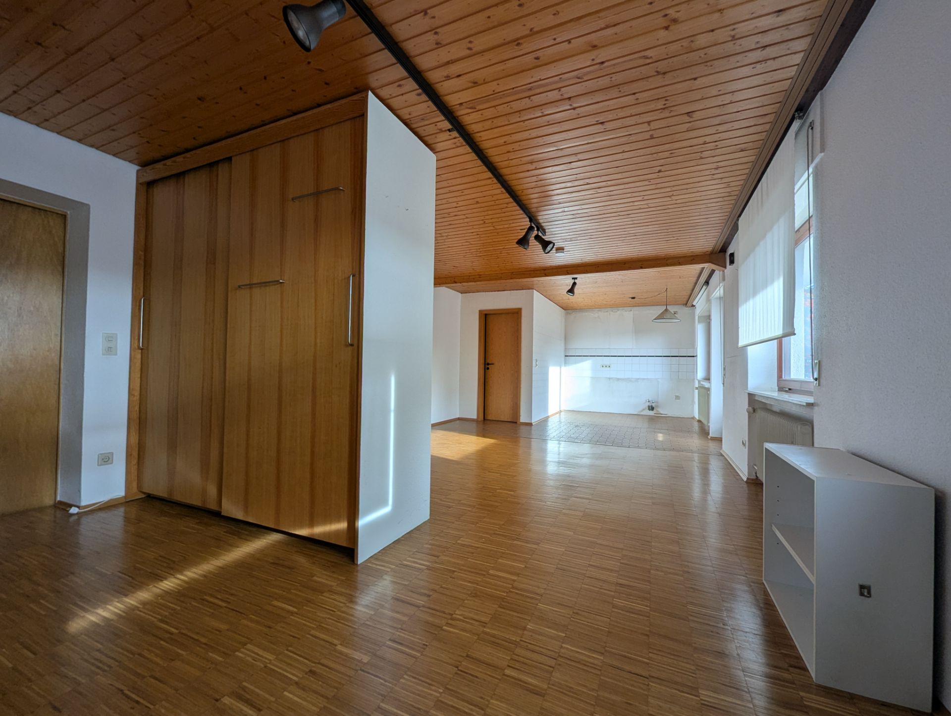 Heller Raum mit Holzboden, Holzdecke, Einbauschrank und Fenstern, offene Küche im Hintergrund.