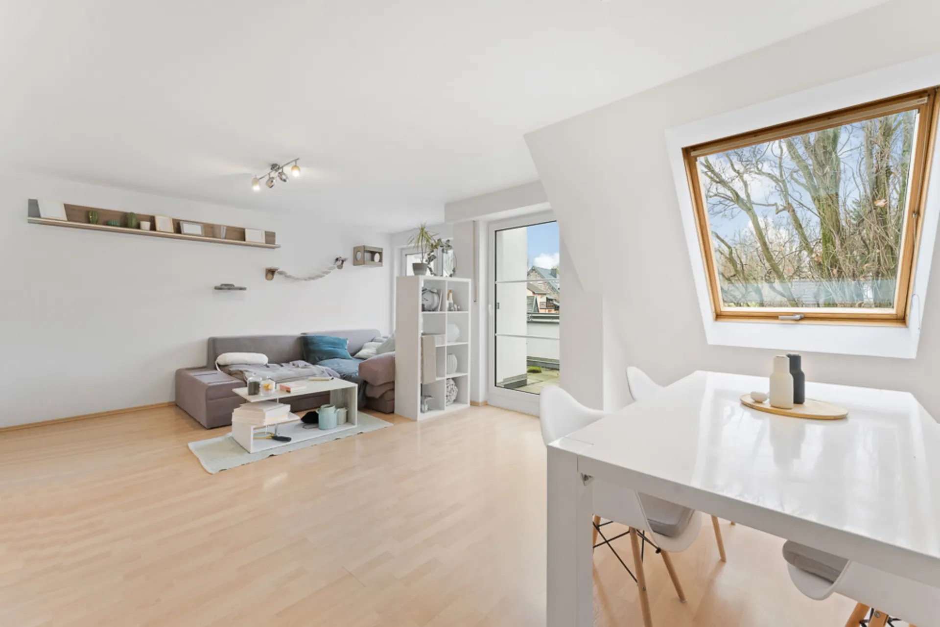 Helles Wohnzimmer mit Sofa, Esstisch, Regal und großem Fenster mit Blick nach draußen.