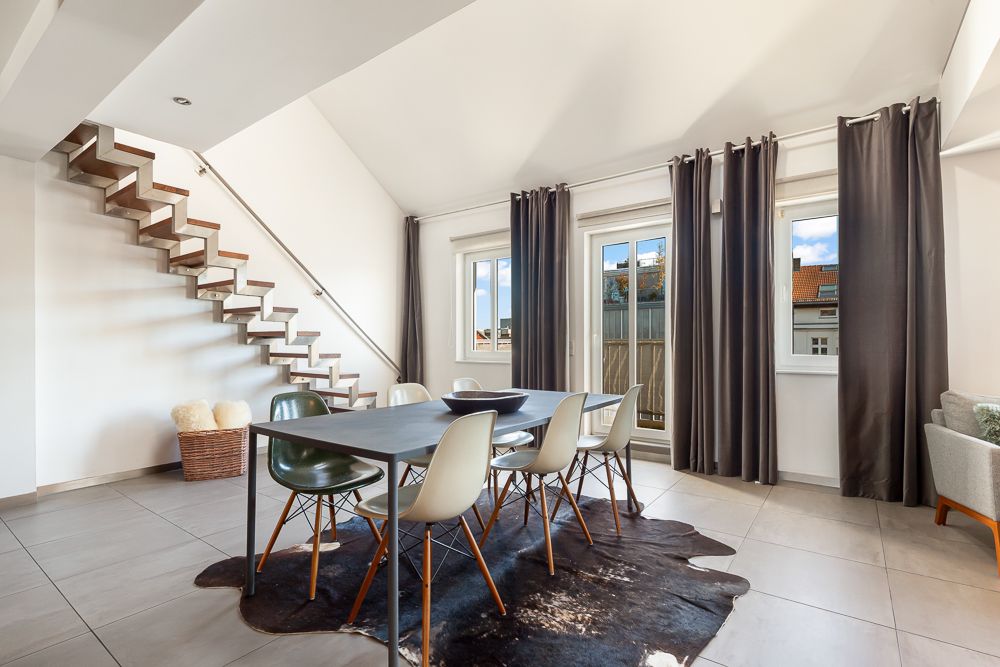Heller Essbereich mit Tisch, sechs Stühlen und Teppich, daneben moderne Treppe und große Fenster.