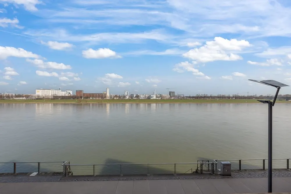 Blick auf einen breiten Fluss mit einer Stadt und blauem Himmel im Hintergrund.