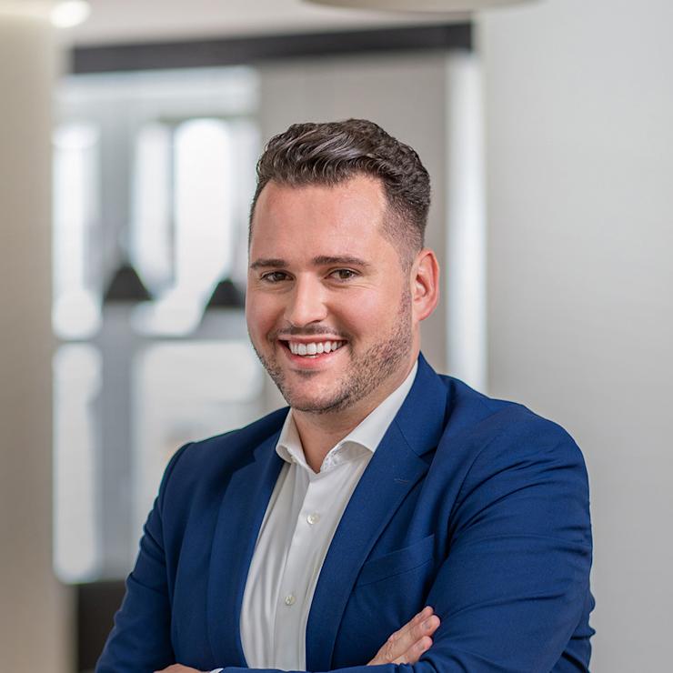 Ein Mann im blauen Anzug steht lächelnd mit verschränkten Armen in einem modernen Innenraum.