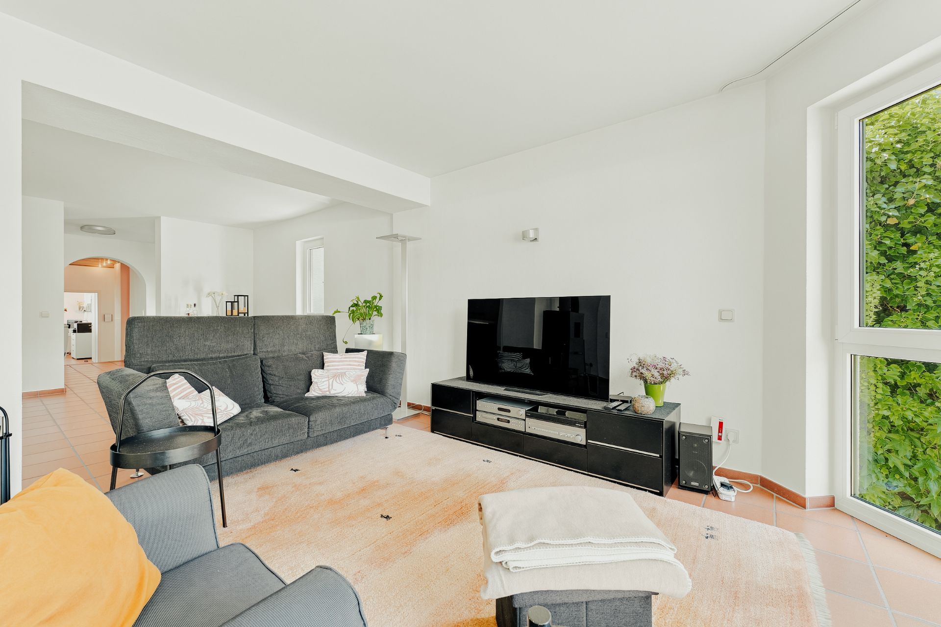 Helles Wohnzimmer mit grauem Sofa, Fernseher, Teppich und großen Fenstern mit Blick ins Grüne.
