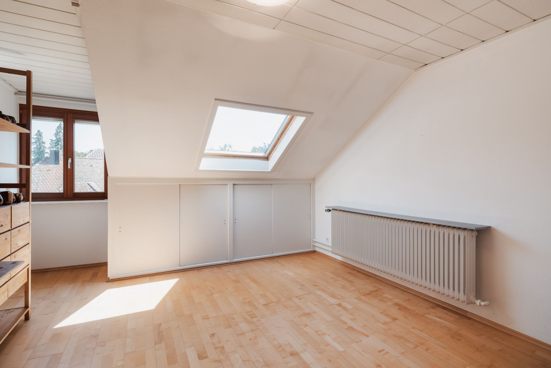 Helles Dachzimmer mit Holzfußboden, Dachfenster, Heizkörper und Einbauschränken unter der Schräge.