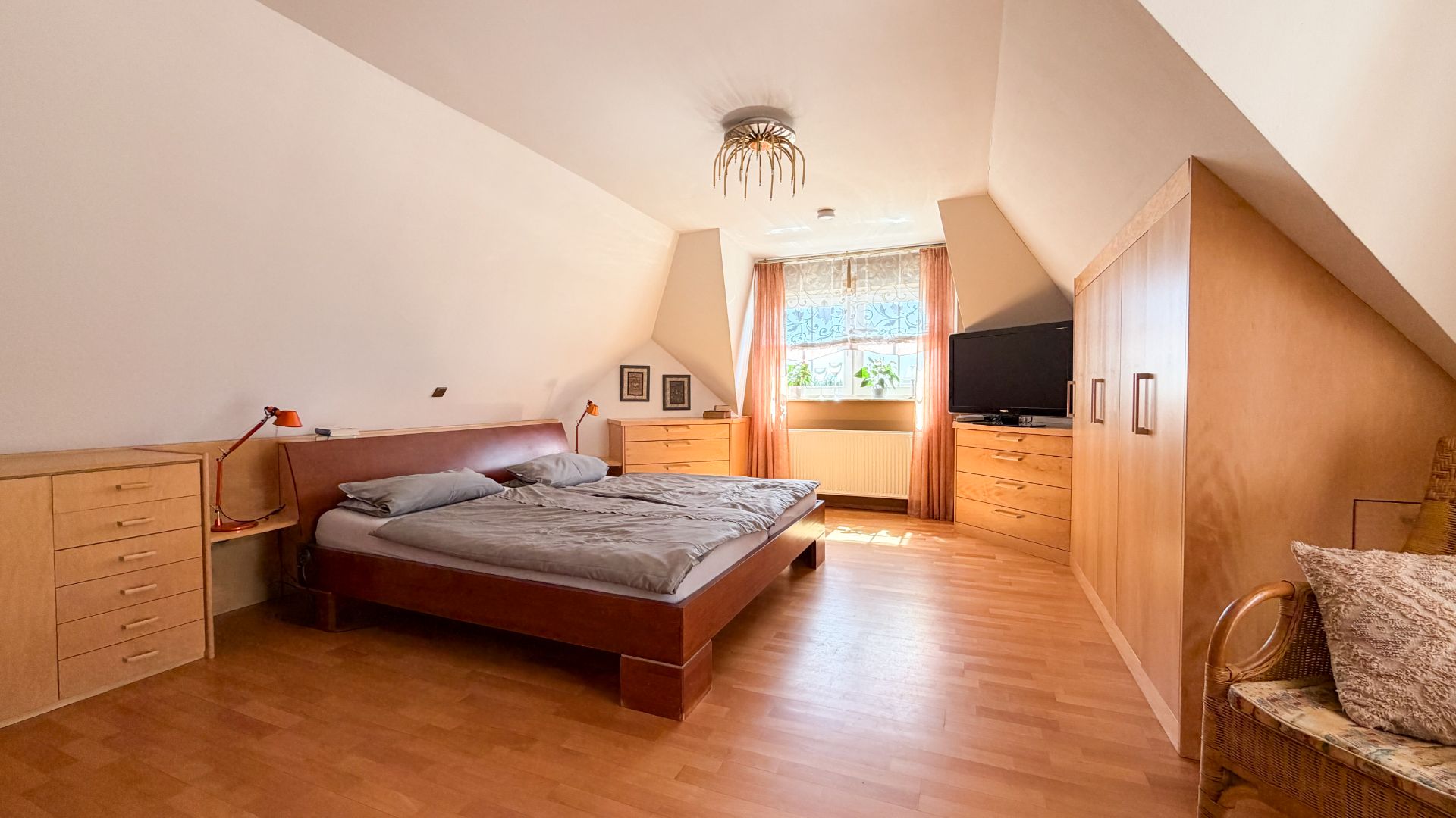 Helles Schlafzimmer mit Doppelbett, Holzmöbeln und Fenster mit Vorhang.
