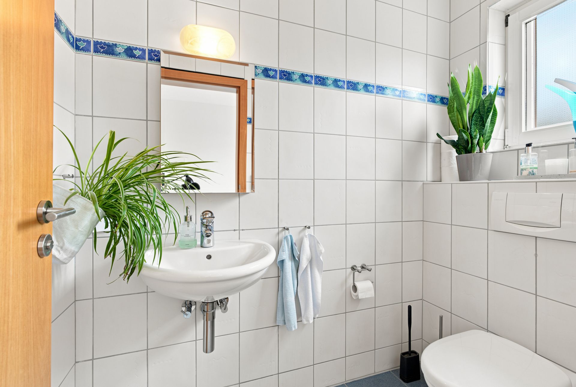 Helles Badezimmer mit Waschbecken, Spiegel, WC und zwei grünen Pflanzen auf Fliesen.
