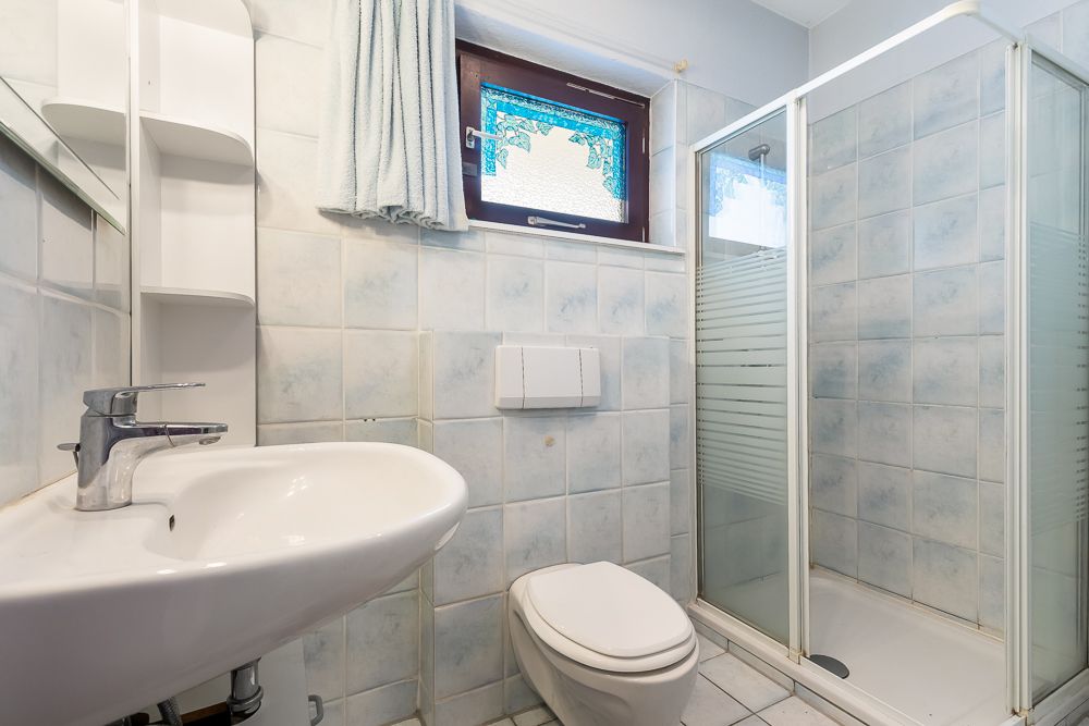Kleines Badezimmer mit Waschbecken, Toilette, Dusche und Fenster mit Vorhang.